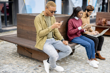 Multiracial students using gadgets outdoors. Concept of education. Remote and e-learning. Idea of students lifestyle. Young guys and girl sitting on wooden bench at university campus