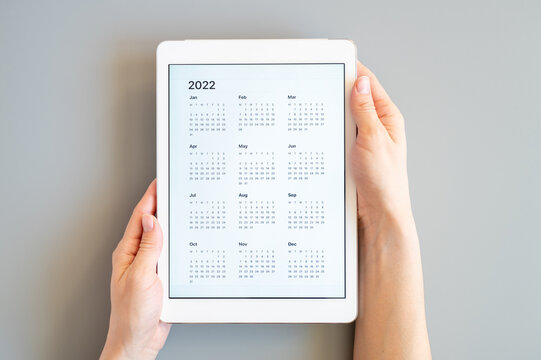 tablet computer with an open app of calendar for 2022 year in a womans hands on a gray background. concept business or to do list goals with technology using. top view, flat lay