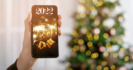 Woman hand holding smartphone with Happy new year 2022 word on screen with light bokeh background. Mockup with space for text.