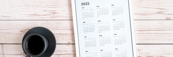 tablet computer with an open app of calendar for 2022 year and cup of tea or coffee on wooden boards background. concept business or to do list goals with technology using. top view, flat lay. banner