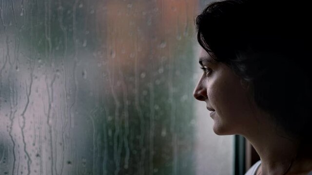 Woman standing by window during rainy day looking outside watching rain