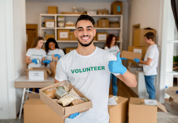 Happy arab man at volunteer center holding donations paper box, showing thumb up and smiling at camera