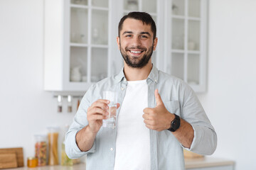 Smiling handsome adult caucasian man hold glass with water and showing thumb up