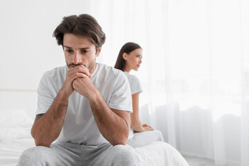 Depressed young caucasian husband with stubble sits on bed and sad, ignoring offended lady in...