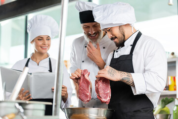 Asian chef holding raw meat near smiling colleagues with cookbook in kitchen