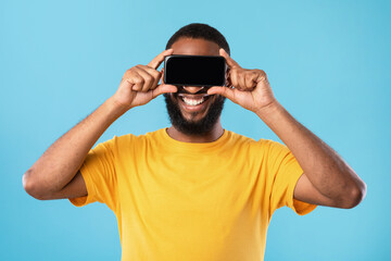 Fototapeta premium Happy black guy covering eyes with blank smartphone, ofering mockup for website or application on blue studio background
