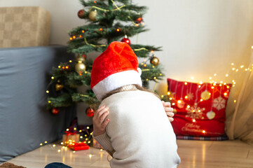 Little girl in Santa hat sitting with back turned on the background of decorated Christmas tree disappointed with gift. Cute little child feeling unhappy with bad holiday present