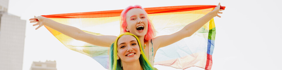 Young lesbian couple laughing while posing with rainbow flag