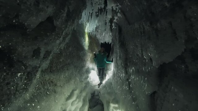 Gimbal approach to digger tourist inside night winter narrow cave, frozen icicles splashes of sharp icebergs. People Illuminates mystical grotto with flashlight. Cold arctic season miracle of nature