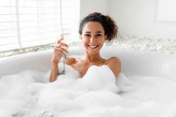 Happy young woman drinking champagne while lying in hot bubble bath near window at home
