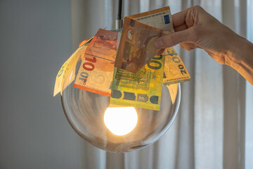 Euro bills over glowing lamp and a woman hand holding Euro banknote. Electricity cost, electricity...