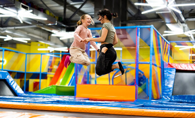 Pretty girls jumping together on colorful trampoline at playground park. Two sisters having fun during active entertaiments indoor - obrazy, fototapety, plakaty