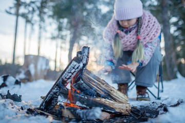The girl in winter clothes warms her hands by the fire in snow-covered forest, winter trip at weekend. Family winter picnic, active lifestyle, focus on woods