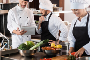 Smiling chef holding cookbook near asian colleague and vegetables in kitchen