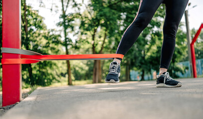 Young girl woman exercising outdoors with rubber elastic band doing training for her legs wearing black leggings. Closeup view of active workout with additional sport equipment outside