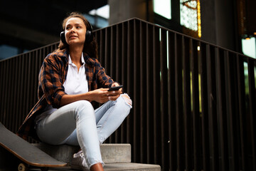 Beautiful young girl with skateboard. Urban brunette woman listening the music outside.