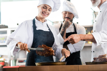 Chef pointing at roasted steak near multiethnic colleagues in aprons in kitchen