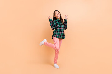Fototapeta na wymiar Photo of excited funny school girl dressed checkered clothes smiling walking showing tow v-signs isolated beige color background
