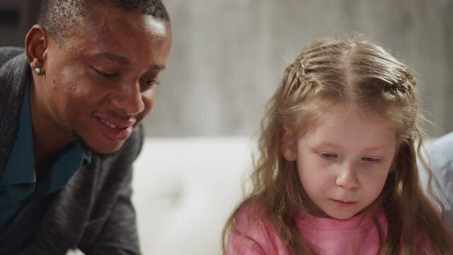 Smiling African-American teacher helps cute little girl do English language homework at private tuition with native speaker closeup slow motion