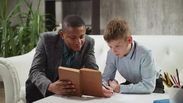 Black private tutor reads book with positive junior schoolboy studying English language at home lesson with native speaker in stylish apartment slow motion