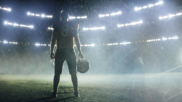 American Football Staduim: Lonely Athlete Warrior Standing on a Field Holds his Helmet and Ready to Play. Player Preparing to Run, Attack and Score Touchdown. Rainy Night with Dramatic Fog - Powered by Adobe