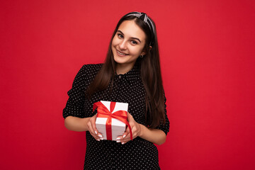 Beautiful happy brunette girl isolated over colourful background wall wearing stylish casual clothes holding gift box and looking at camera