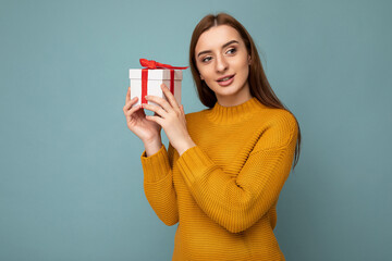 Photo shot of beautiful happy brunette young woman isolated over blue background wall wearing mustard sweater holding white gift box with red ribbon and looking to the side