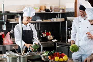 Smiling chef washing broccoli near interracial colleagues talking in restaurant kitchen