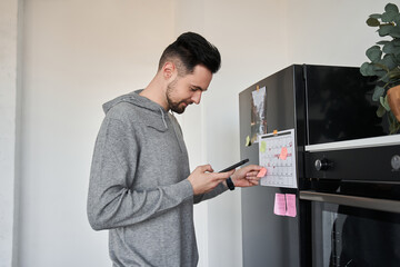 Naklejka na ściany i meble Man looking at the smartphone and putting sticker with notification at the refrigerator