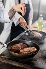 Cropped view of tattooed chef pouring salt while roasting meat on cooktop in kitchen