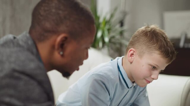 Young black man tutor conducts private English lesson to primary schoolboy at homeschooling with native speaker in living room closeup slow motion