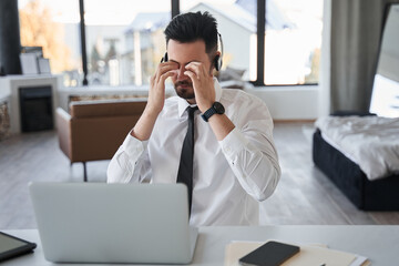 Bearded man wearing headset rubbing his eyes while sitting in front of the laptop screen