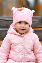 Portrait of a child girl sitting on a bench in autumn city park. Beautiful nature, trees with yellow leaves.