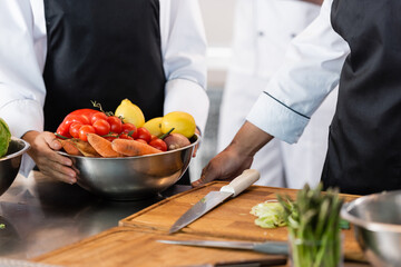 Cropped view of chef holding bowl with ripe vegetables near colleague and chopping boards in kitchen