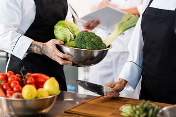 Cropped view of tattooed chef holding bowl with vegetables near colleague with knife in kitchen