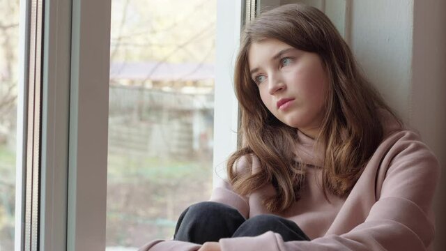 Sad girl looks out the window while sitting on the windowsill. Depressive state in a teenager.