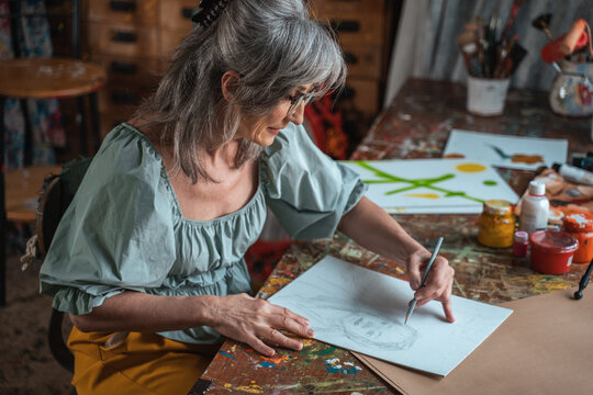 Grey haired woman in casual clothes sitting at the table of her art studio and drawing