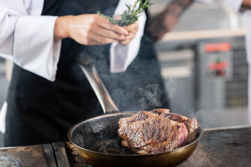 Roasted meat on frying pan near blurred chef holding rosemary in kitchen