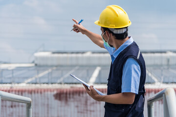 Worker pointing forward with clipboard in hand at factory building background