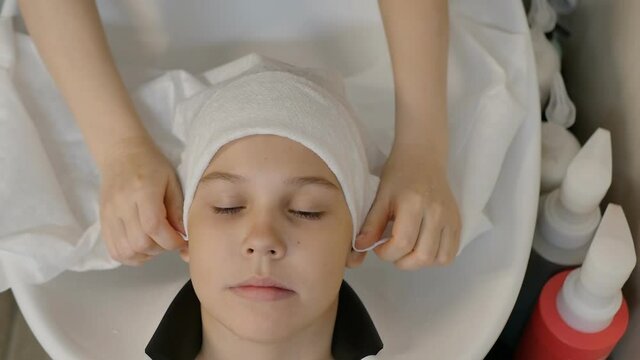 top view, slow motion: hairdresser covers teenage girl's hair with white towel after shampooing before hair styling. Cute face of a Caucasian girl in a white headscarf