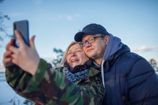 Reunion of couple on the shore of beautiful frozen lake in winter. Middle aged man and woman in military jacket with camouflage pattern look at smartphone camera to make selfie