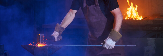 Professional mature blacksmith in safety apron and gloves beating on molten metal with hammer for...
