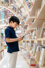 Young Asia boy standing in the modern and nice interior bookstore. Choosing and holding a book. Many bookshelf in the shop. Kid learning and education concept. Colorful book at library