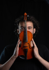 Portrait of a young dark-haired man with a beard, looking out from behind an old violin. Dark background. Selective focus.
