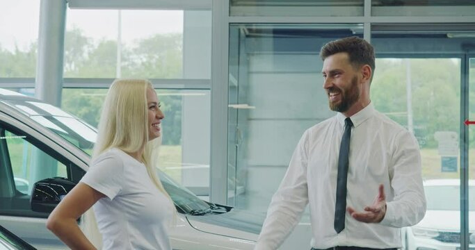 Beautiful woman with blond hair standing at modern salon with luxury car and talking with competent dealer. Bearded salesman helping female client to choose transport at showroom.
