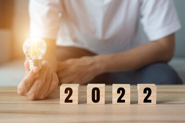A young businessman holding a light bulb in his hand with the number 2022 on a wooden block placed...