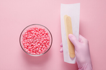 Pink granules of wax for depilation in a glass bowl on a pink background and a hand of a master in a pink glove holding a strip for depilation and wooden spatulas. Epilation, depilation - obrazy, fototapety, plakaty