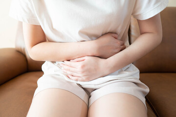 Fototapeta na wymiar Young female suffering form stomach ache while sitting on couch at home. Causes of abdominal pain include menstruation pain, gastritis, stomach ulcer, food poisoning, diarrhea or IBS.