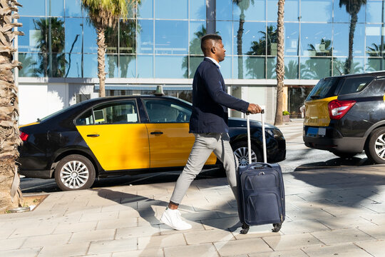 Black businessman with suitcase walking on sidewalk past taxi cars