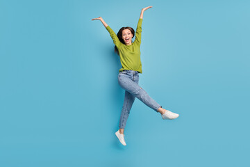 Photo of surprised crazy funky lady jump raise hands wear green shirt jeans sneakers isolated blue...